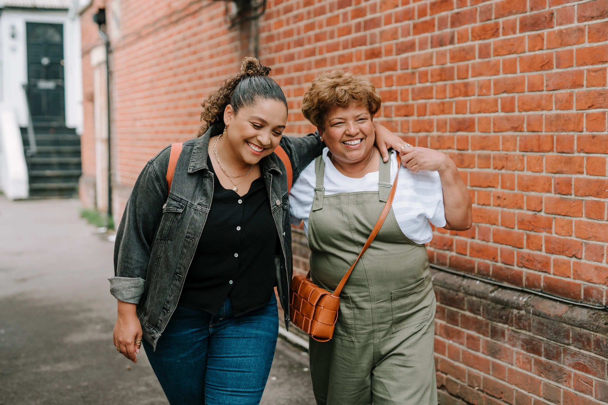 Two smiling adults walking on the sidewalk in front of a brick wall, one person with arm around the other.