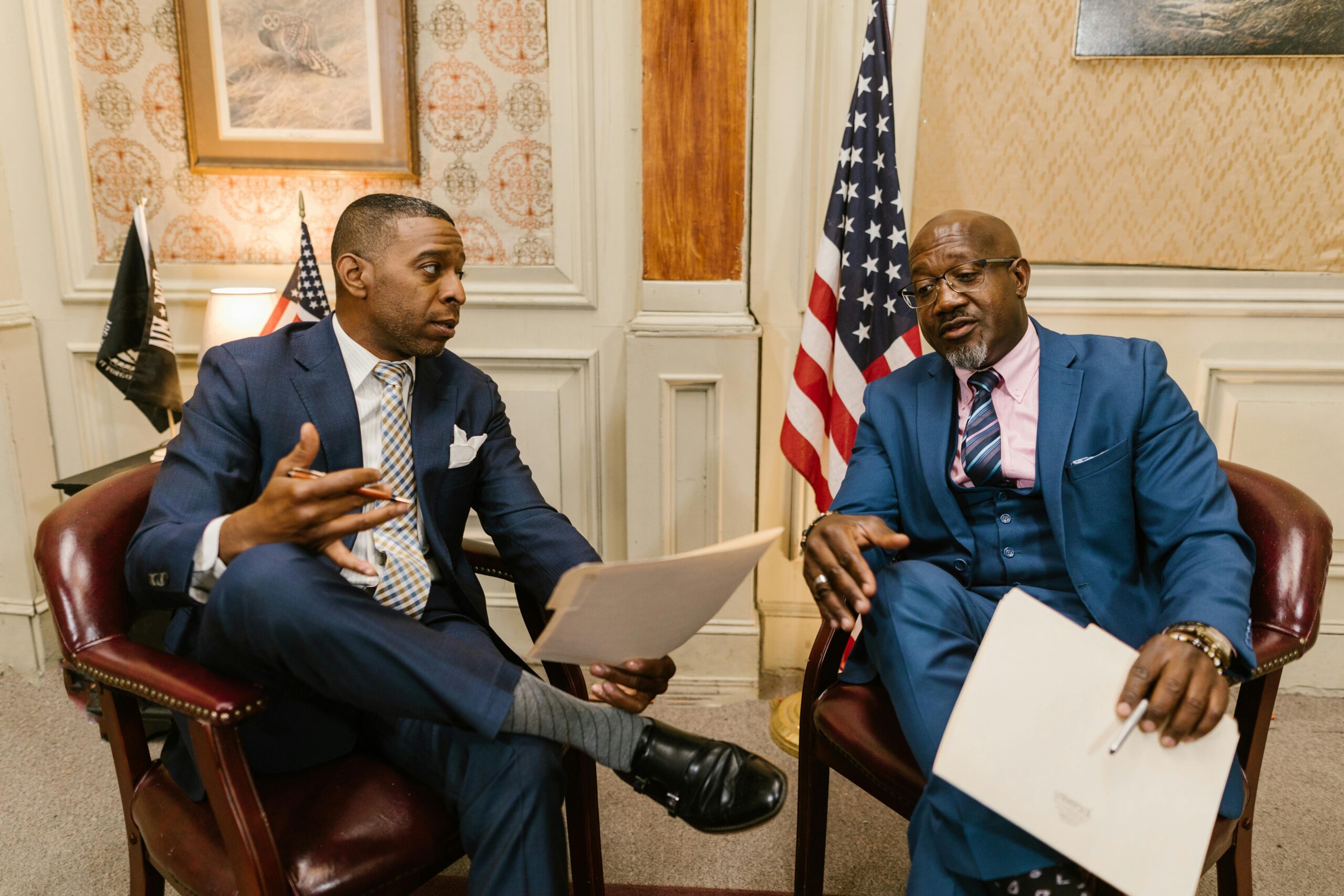 Two men wearing suits have a discussion while sitting in chairs in a carpeted room with an American flag behind them.